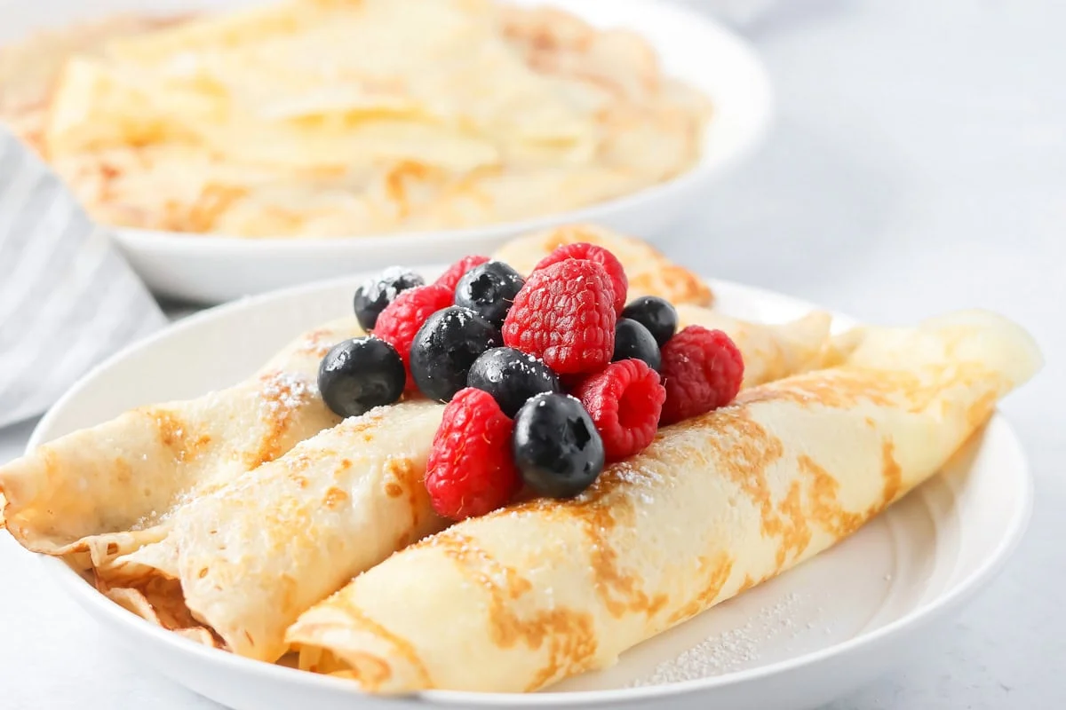 Swedish pancakes rolled and topped with berries on white plate.