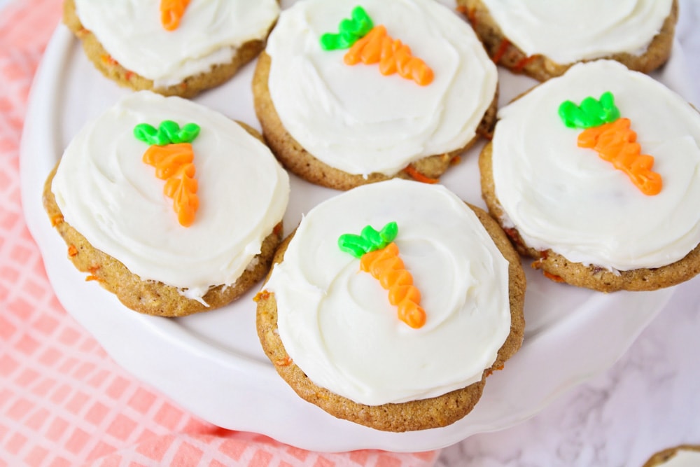 Carrot cake cookies topped with white frosting and little carrots drawn with icing served on a white platter.