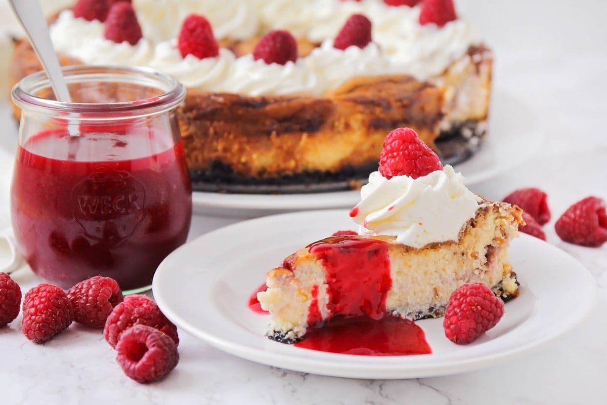 White chocolate raspberry cheesecake on a white plate topped with raspberry sauce.