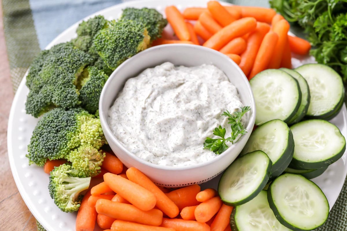 A white bowl of dill vegetable dip surrounded by vegetables.