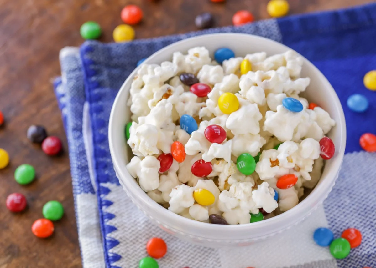 White chocolate M&M popcorn in white bowl with M&Ms sprinkled about.