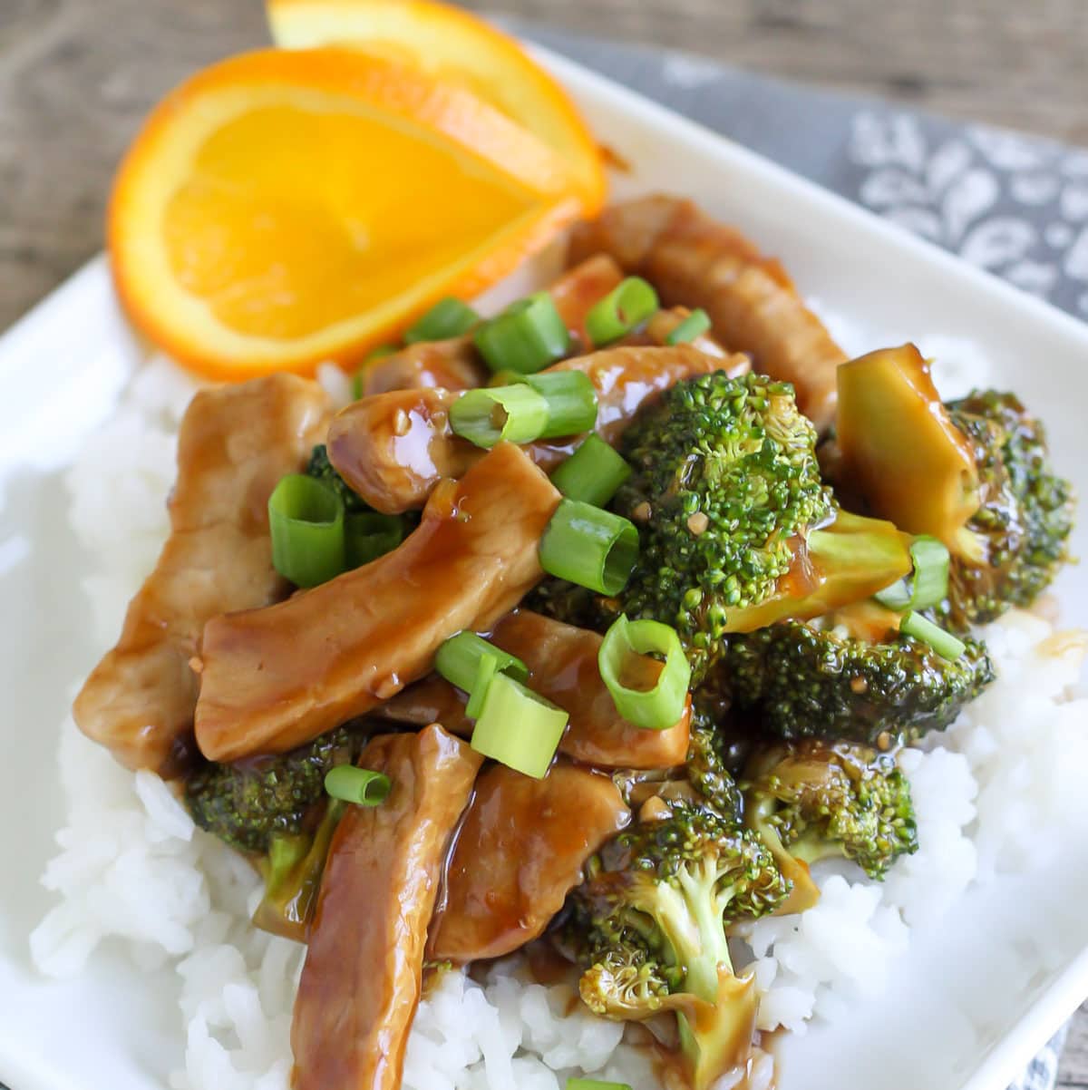 Pork Broccoli Stir Fry garnished with diced green onions and two orange slices on top of white rice on a white plate. 