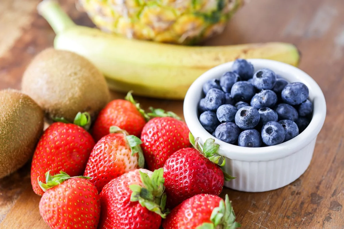 Strawberries, blueberries, kiwis, banana, and pineapple sitting on a wood cutting board.