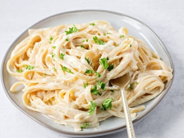 Fettuccine Alfredo recipe on plate with fork in pasta and parsley sprinkled on top.
