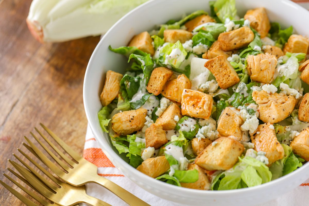 A buffalo chicken green salad in a white bowl with gold forks beside the bowl.  