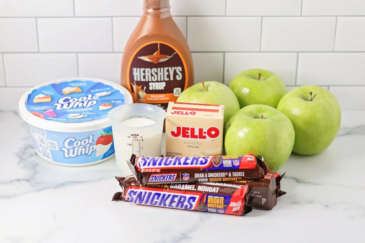 Our 10-minute Snickers salad is a sweet and tart dessert where bright pops of apples perfectly complement chewy chunks of Snickers. Ingredients for Snickers salad on a white countertop.
