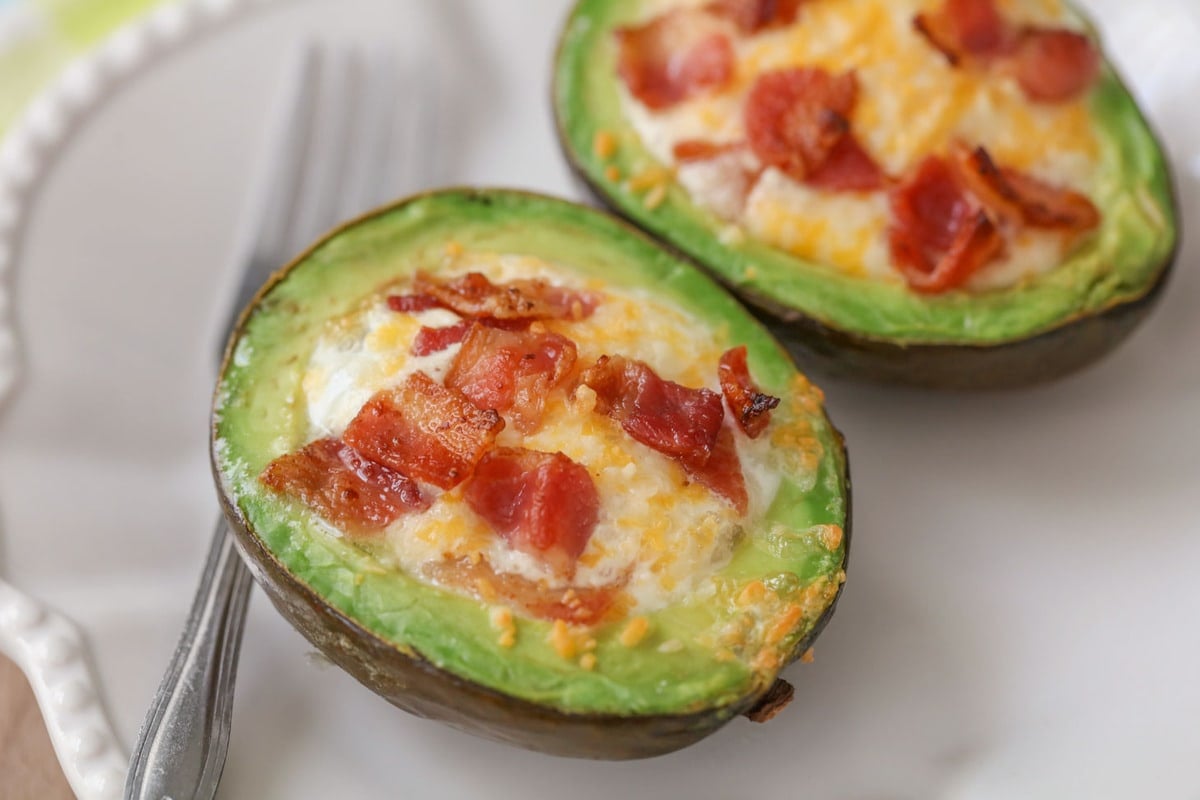 Breakfast Egg Recipes - avocado cut in half with cooked eggs and bacon in each half on a white plate. 