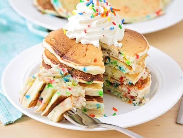 A stack of funfetti pancakes topped with whipped cream and a bite missing.