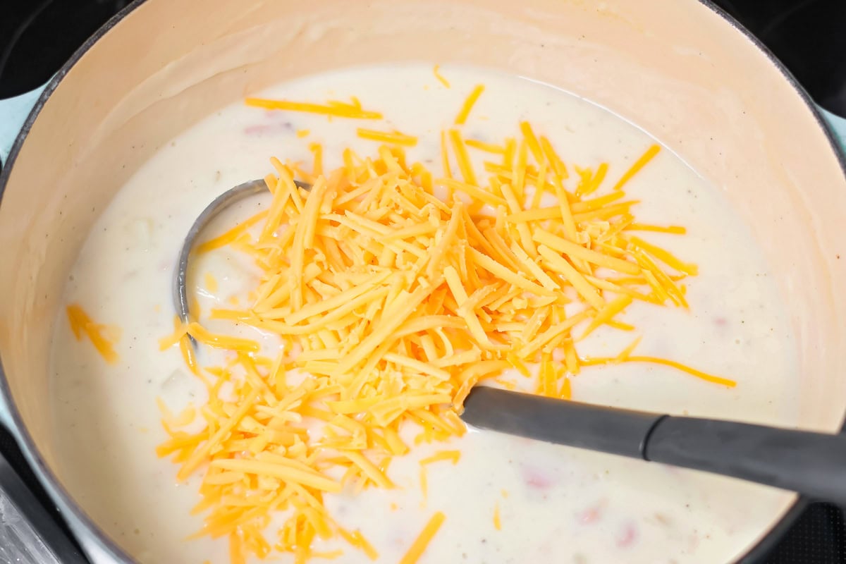 Adding shredded cheese to a pot of ham and potato soup recipe.