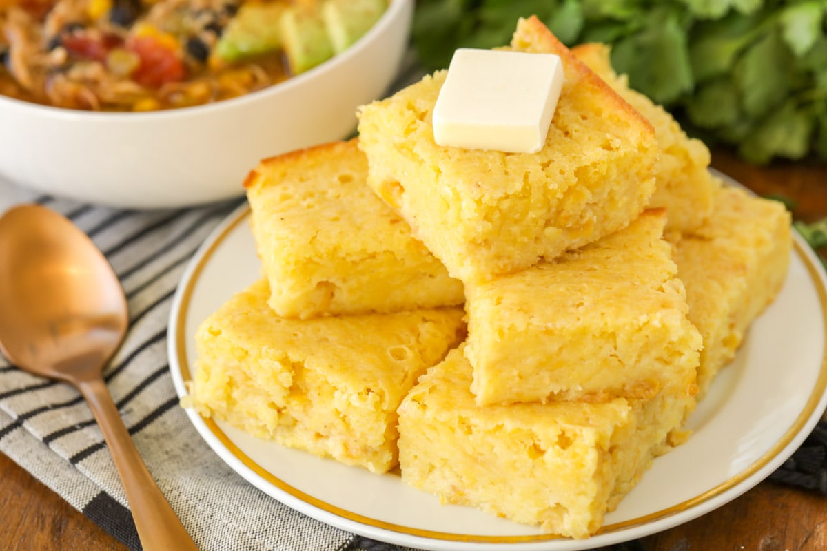 A stack of corn bread on a white gold rimmed plate topped with a pat of butter - a good side dish for fall soups. 