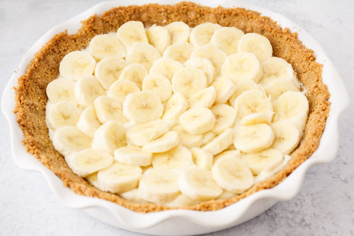 Banana pudding cheesecake filling topped with banana slices in a pie crust in a white pie pan. 