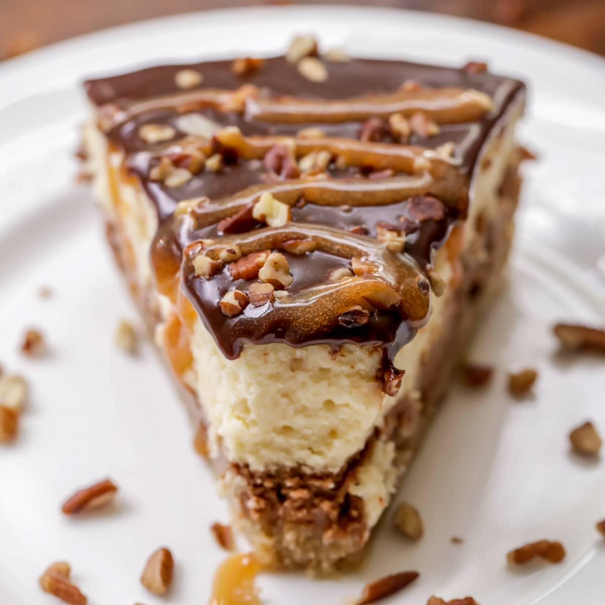 A slice of turtle Cheesecake topped with chocolate, caramel and pecans on a white plate