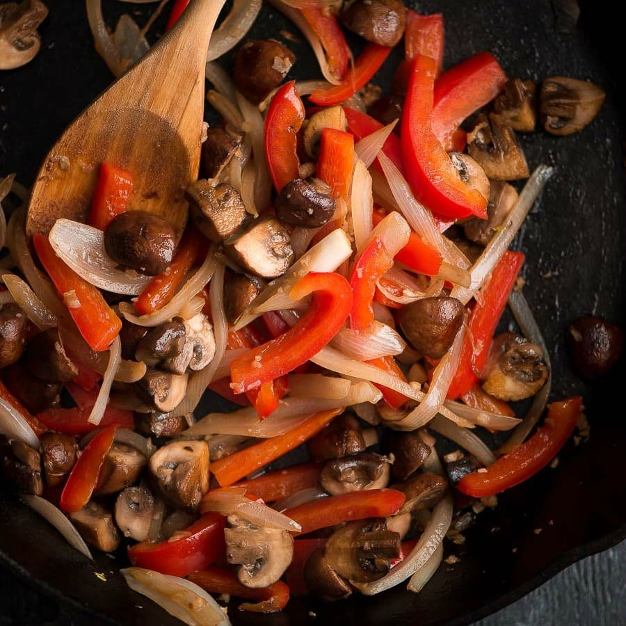 red bell pepper, onions and mushrooms being sautéed in skillet with wooden spoon