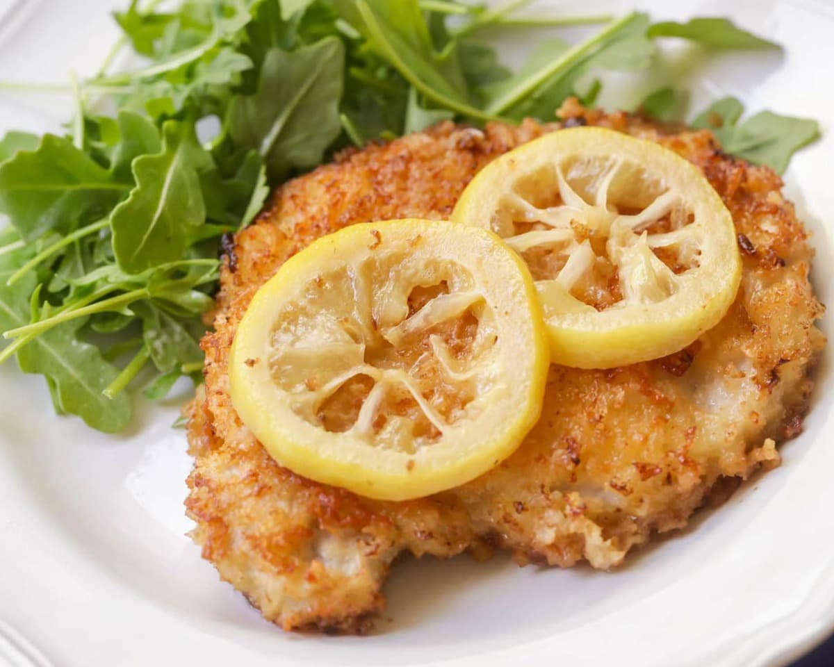 Fried Pork Chops over arugula topped with lemon slices served on a white plate.