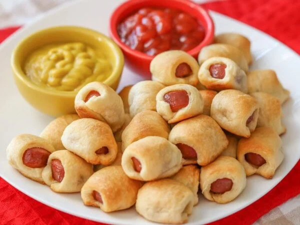 Mini hot dogs stacked on top of white plate with ketchup and mustard.