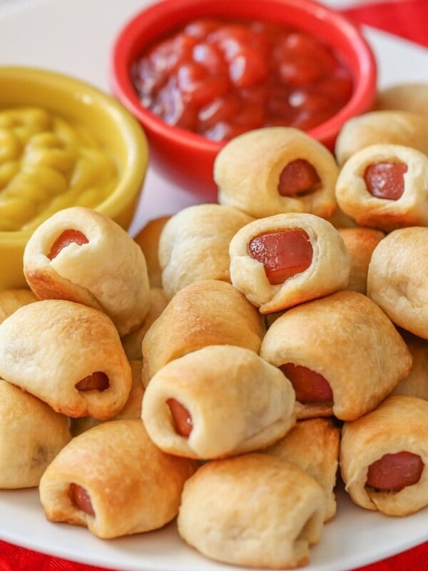 Mini hot dogs stacked on top of white plate with ketchup and mustard.