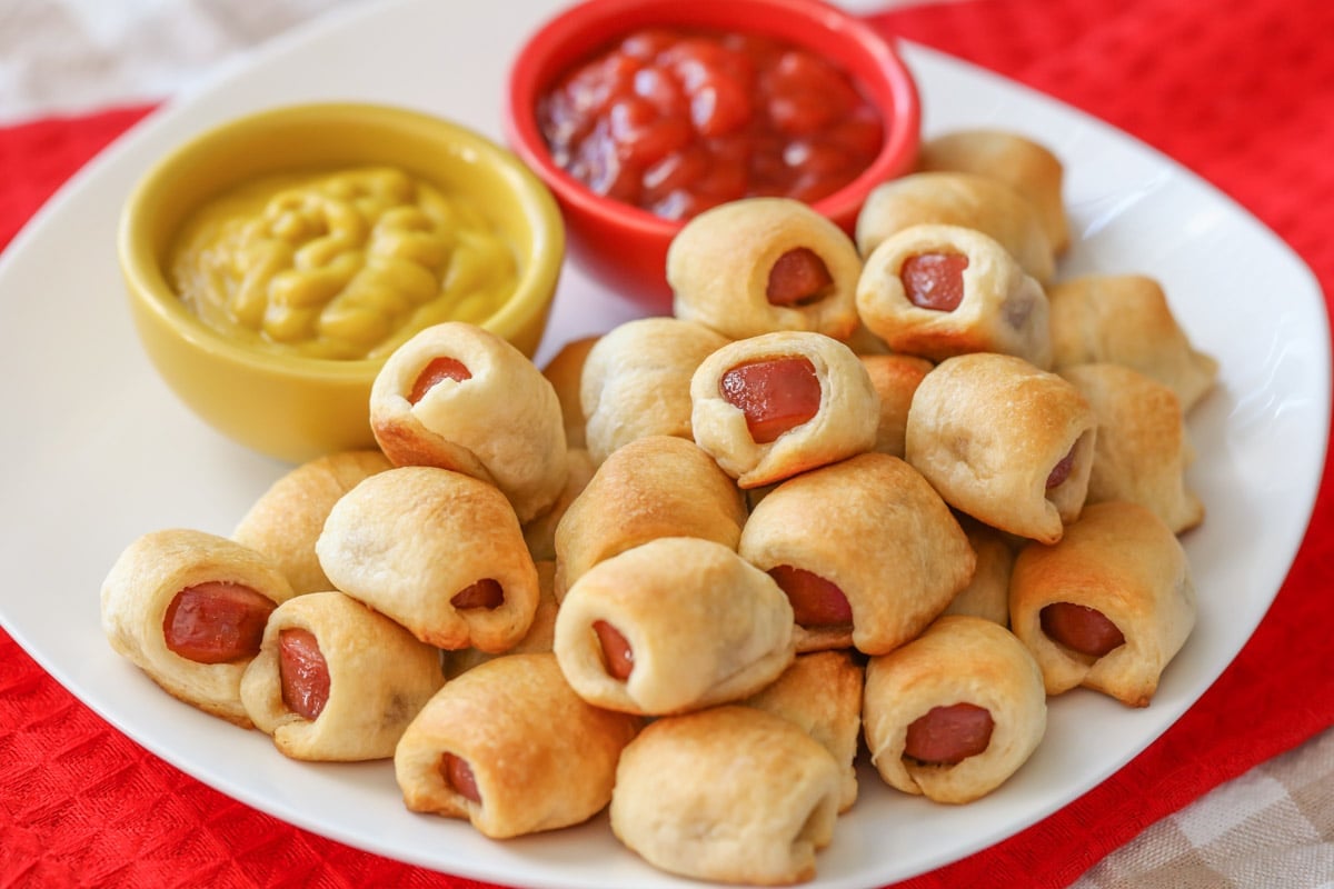 Easy Appetizers - Hot Dog Nuggets on a white plate with a side of ketchup in a small red bowl and a side of mustard in a small yellow bowl. 