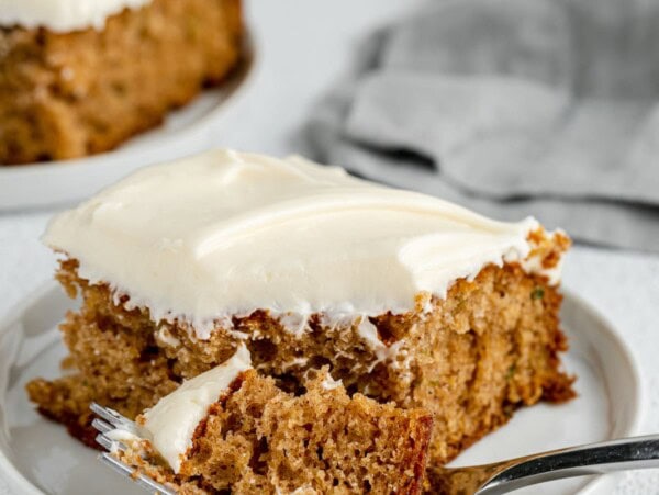 A forkful of zucchini cake next to a frosted cake slice on a white plate.