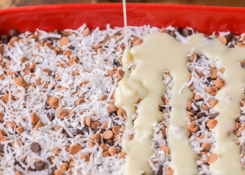 Pouring sweetened condensed milk over layers of cookie and chips.