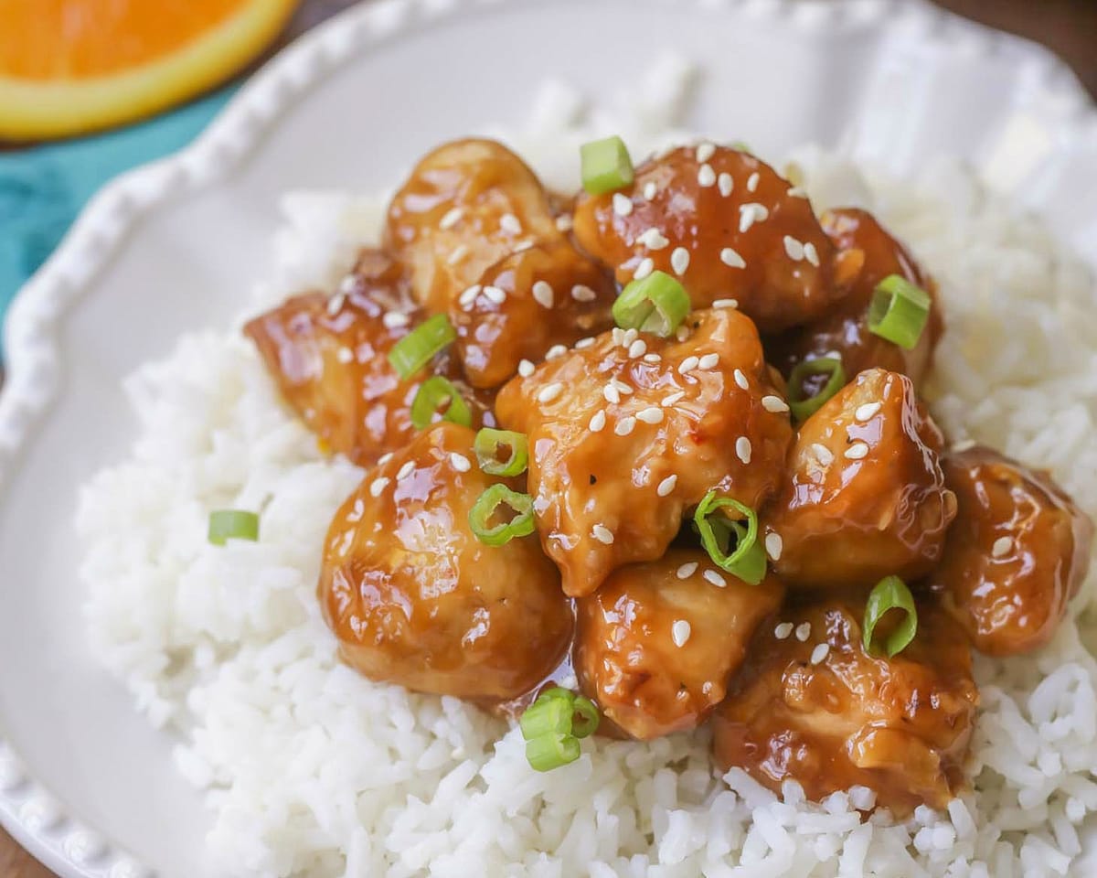 Asian Dinner Recipes - Orange Chicken topped with sesame seeds and diced green onions on top of white rice on a white plate. 