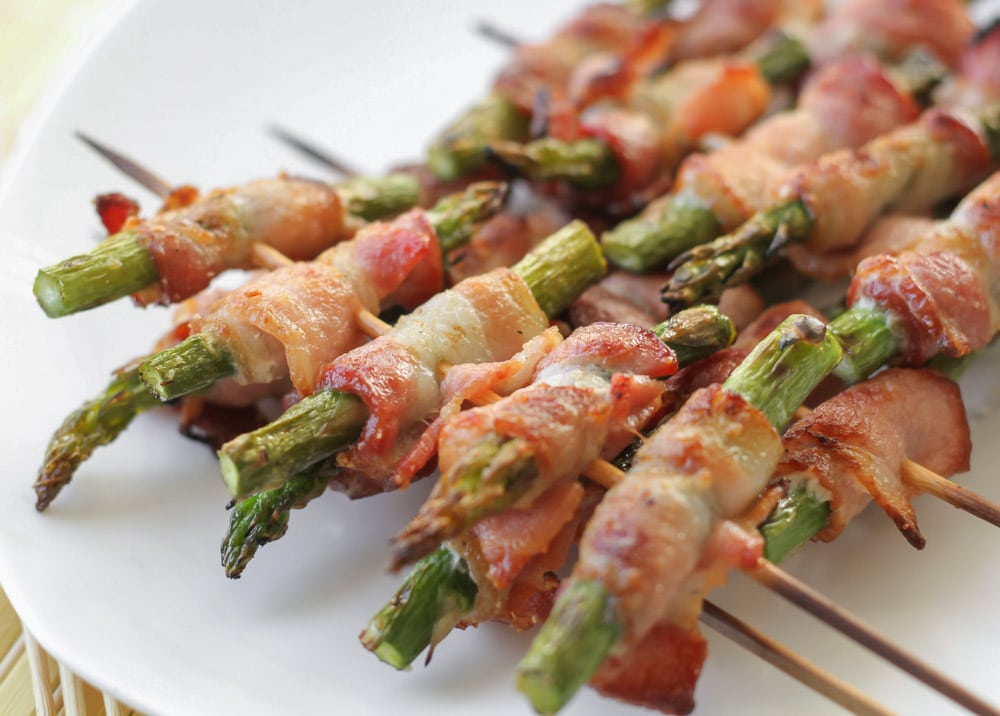 Vegetable side dishes - bacon wrapped asparagus skewers piled on a white plate.