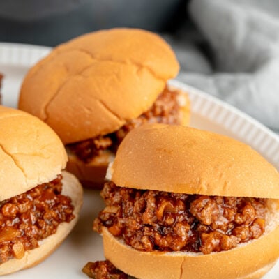 Three sloppy joe sandwiches on a white plate, with a skillet in the background.