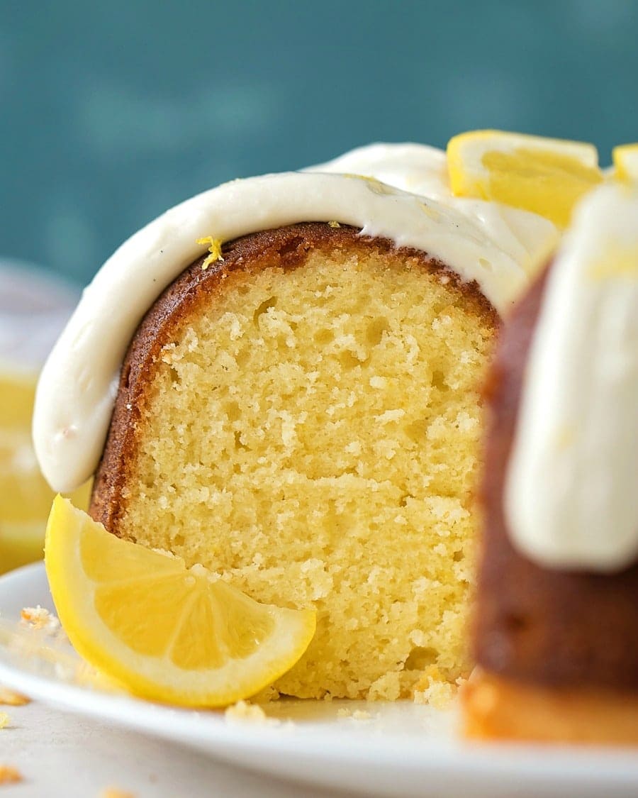 Close up of cream cheese frosting on lemon Bundt cake garnished with a lemon wedge on a white plate. 