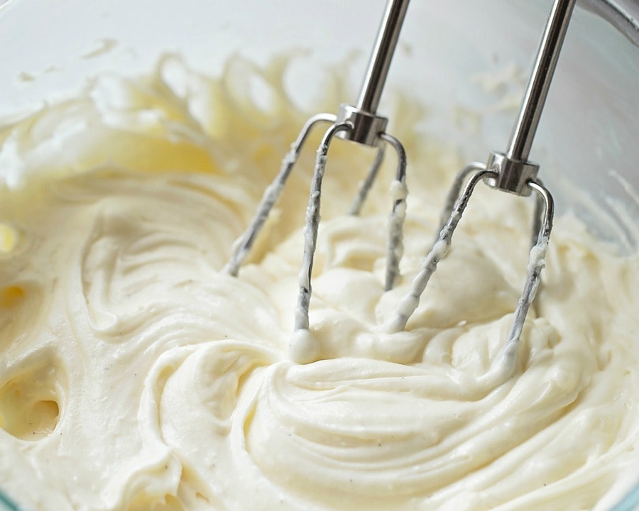 Bundt cake icing being mixed with a hand mixer in a glass bowl. 