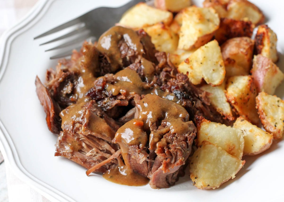 A serving of pot roast topped with gravy on a white plate with a side of potatoes.
