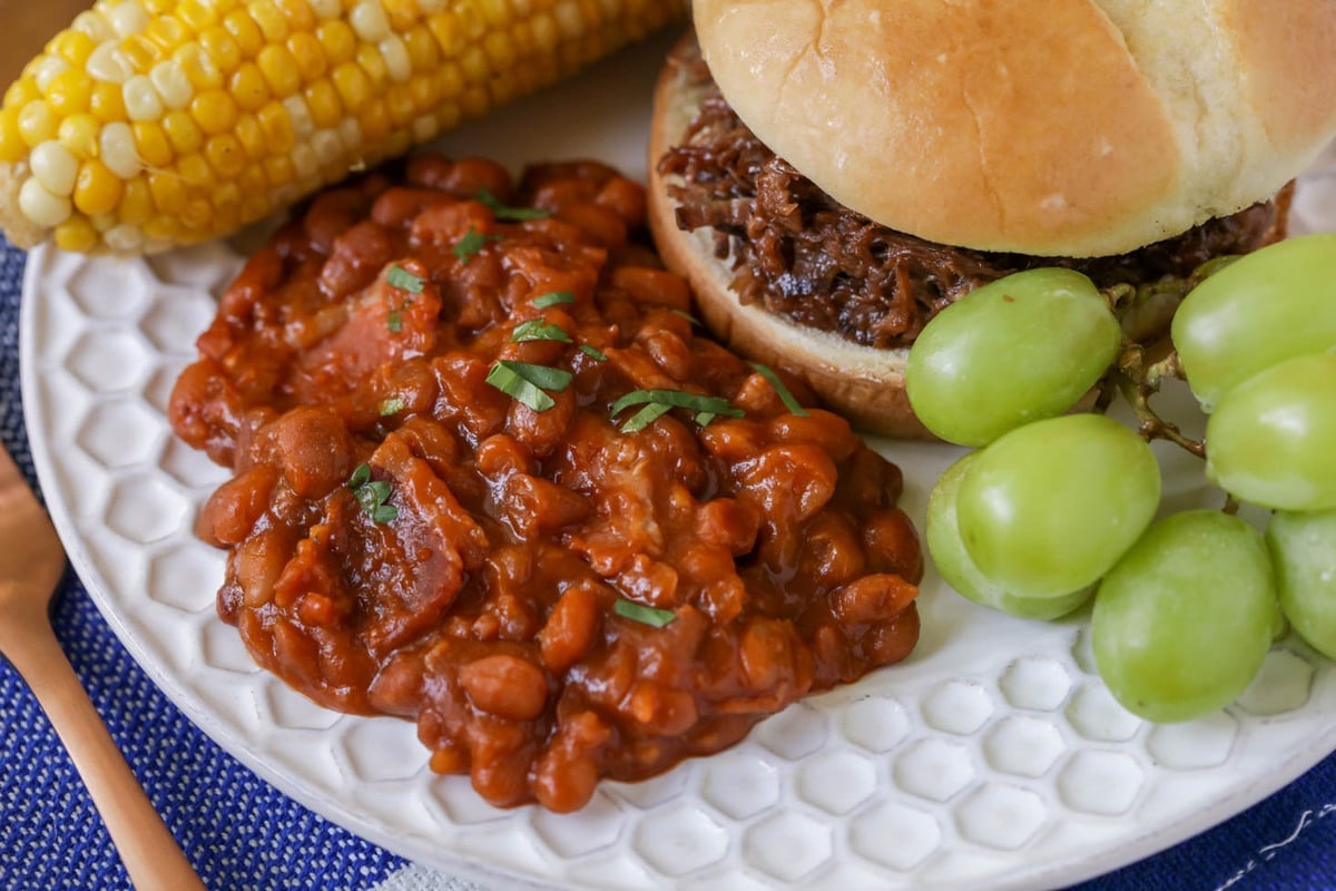 Baked beans on a plate with a pulled pork sandwich, corn on the cob, and grapes.