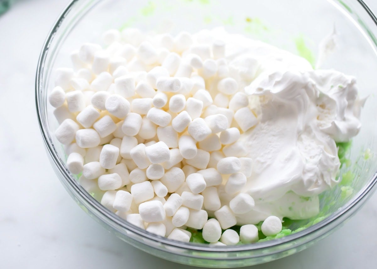 Pudding mix, cool whip, and marshmallows mixed in a white bowl for pistachio salad.