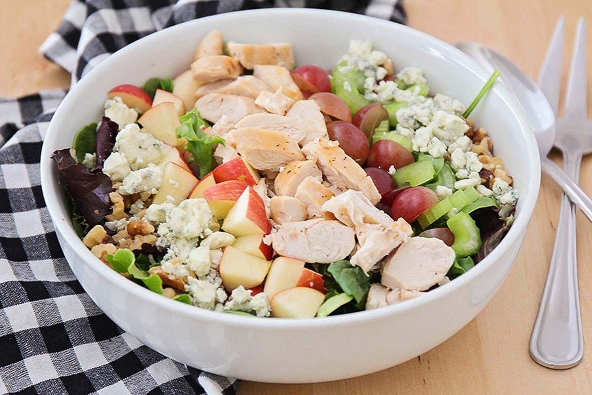 Waldorf salad in a white bowl with a silver serving fork and spoon beside the bowl. 