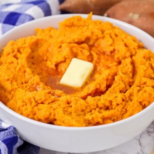Mashed sweet potatoes prepared in white bowl with butter pat on top.