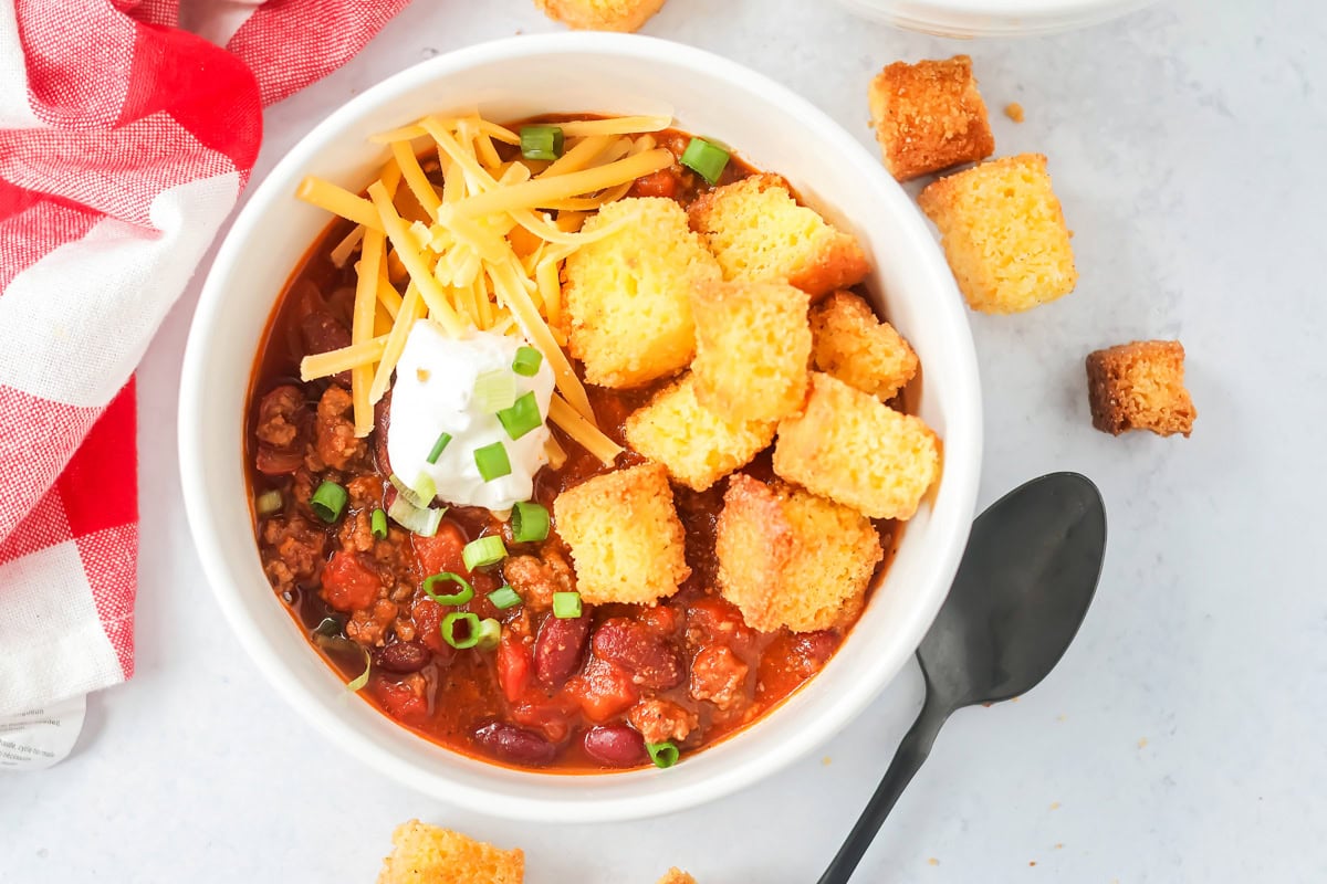 You're going to fall hard for this spicy, hearty homemade beef chili. This Best Chili Recipe is not only simple, but delicious!! Easy chili recipe close up image with cornbread croutons and toppings on top.
