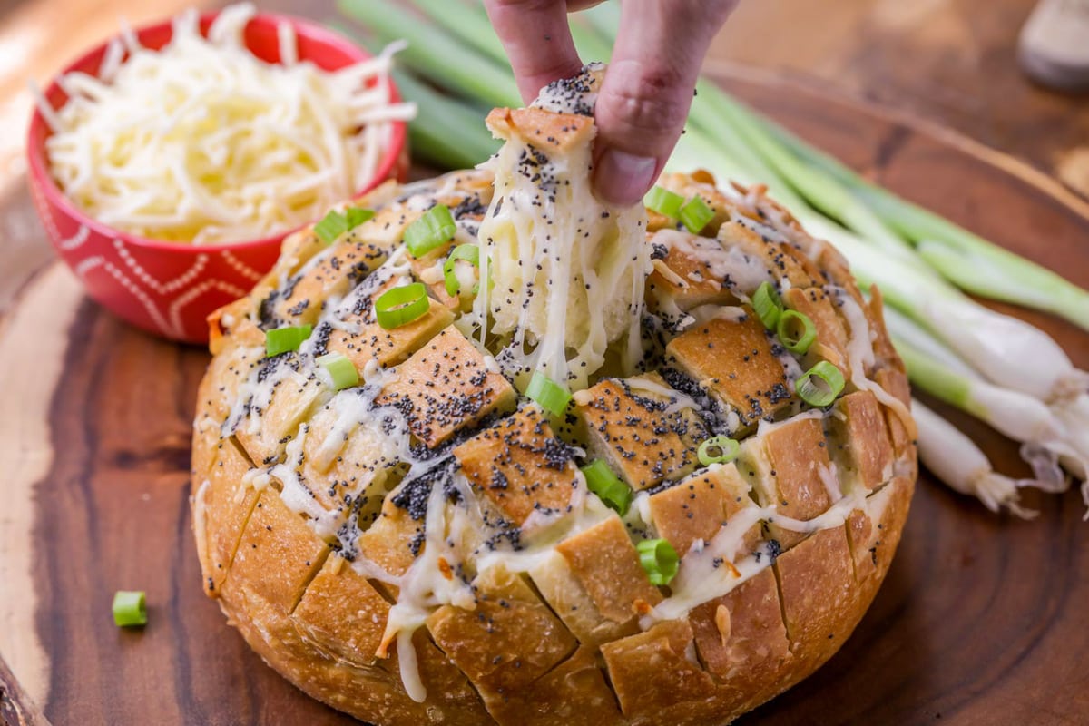  A loaf of pull apart bread with a piece of bread and mozzarella cheese being pulled out of the middle of the loaf with a side of mozzarella cheese in a red bowl on round wooden platter. 