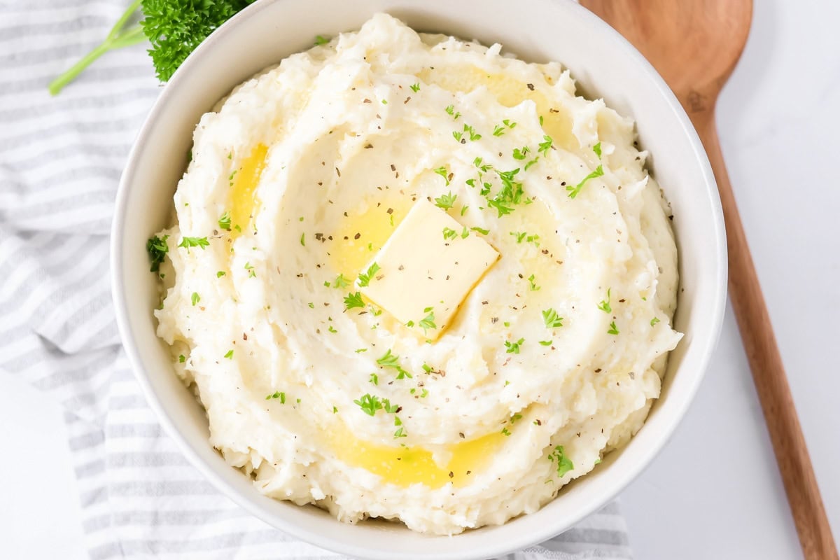 Mashed potatoes recipe served in white bowl with butter, pepper and parsley on top.