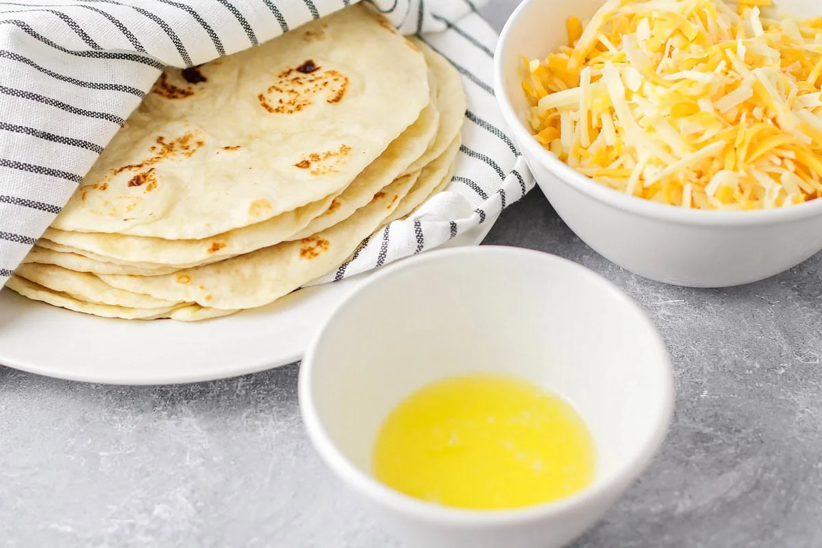 This easy, cheesy chicken quesadilla is a crowd favorite. Filled with a delicious homemade sauce, these hand-held bites are irresistible! Tortillas, cheese, and butter on a kitchen counter.