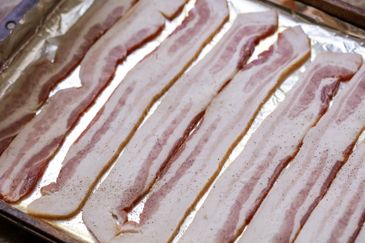 Uncooked bacon slices on a tin foil lined baking sheet.