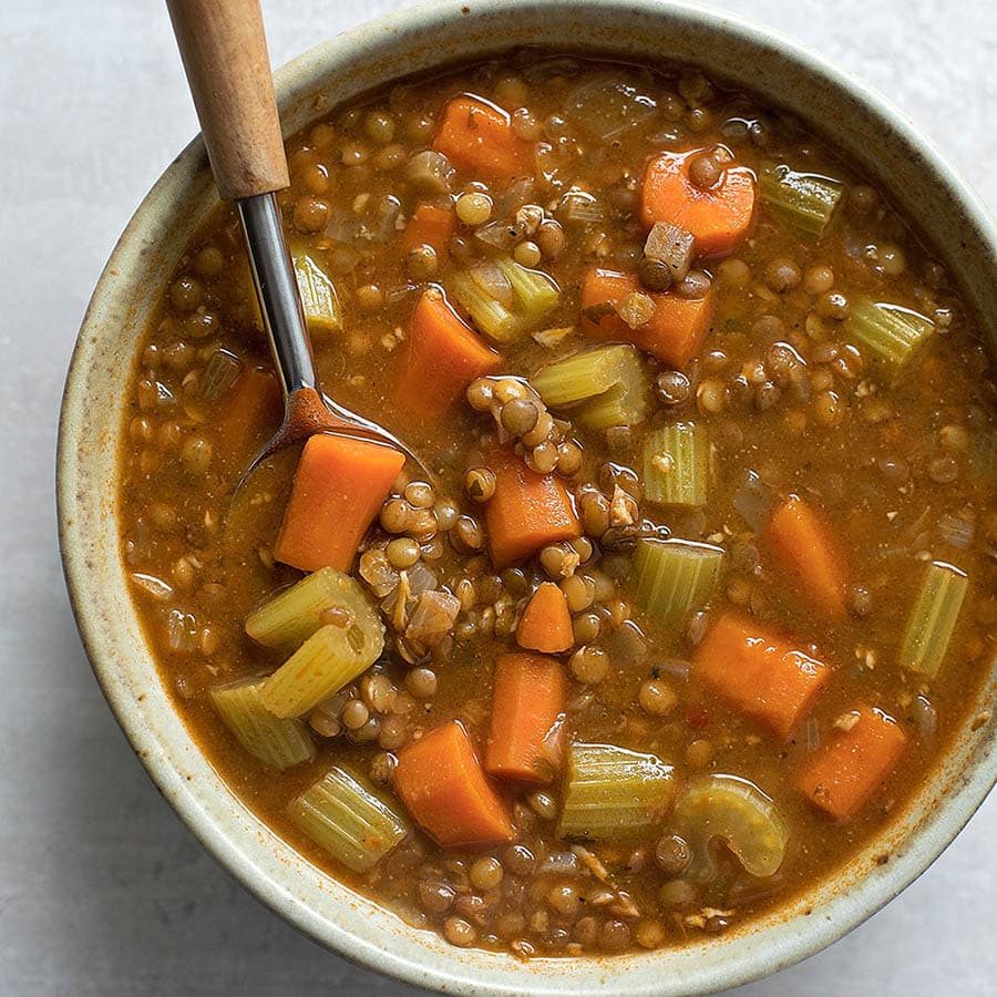 Lentil soup in a tan bowl with a metal spoon with a wooden handle in the bowl.