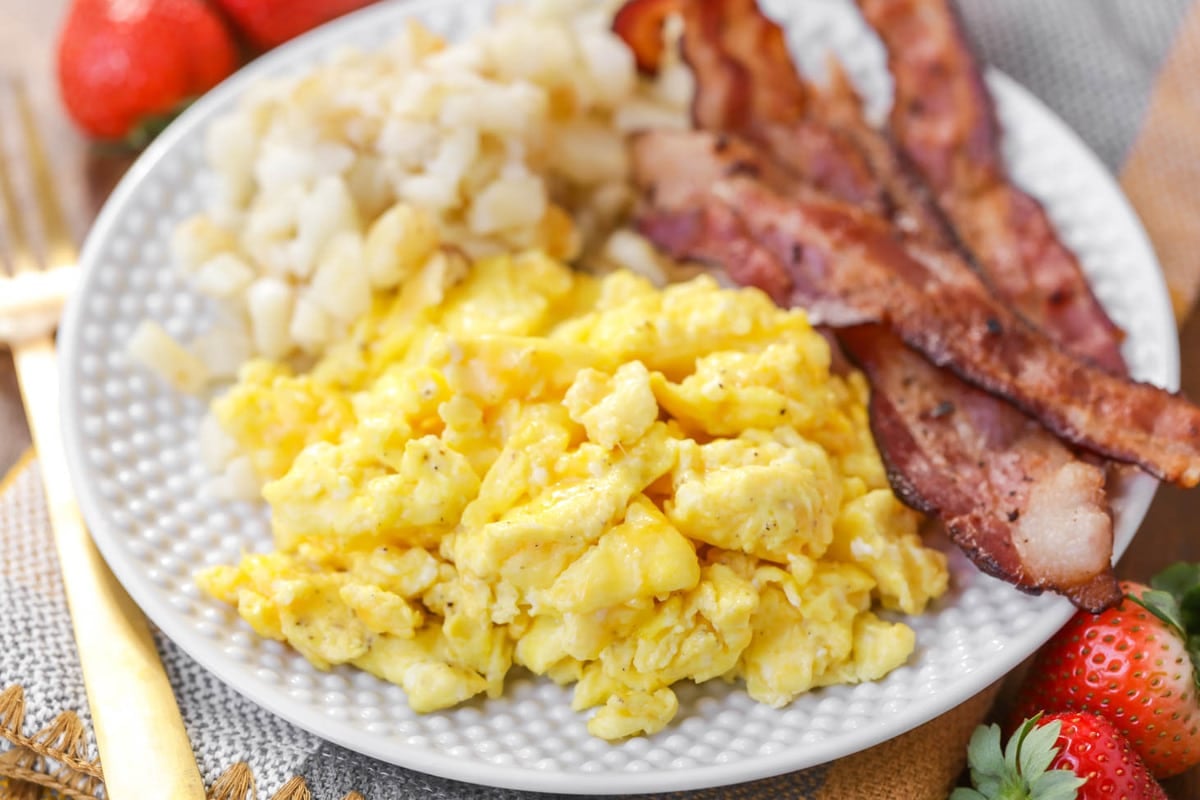 Scrambled eggs with sides of bacon and diced potatoes on a white plate. 