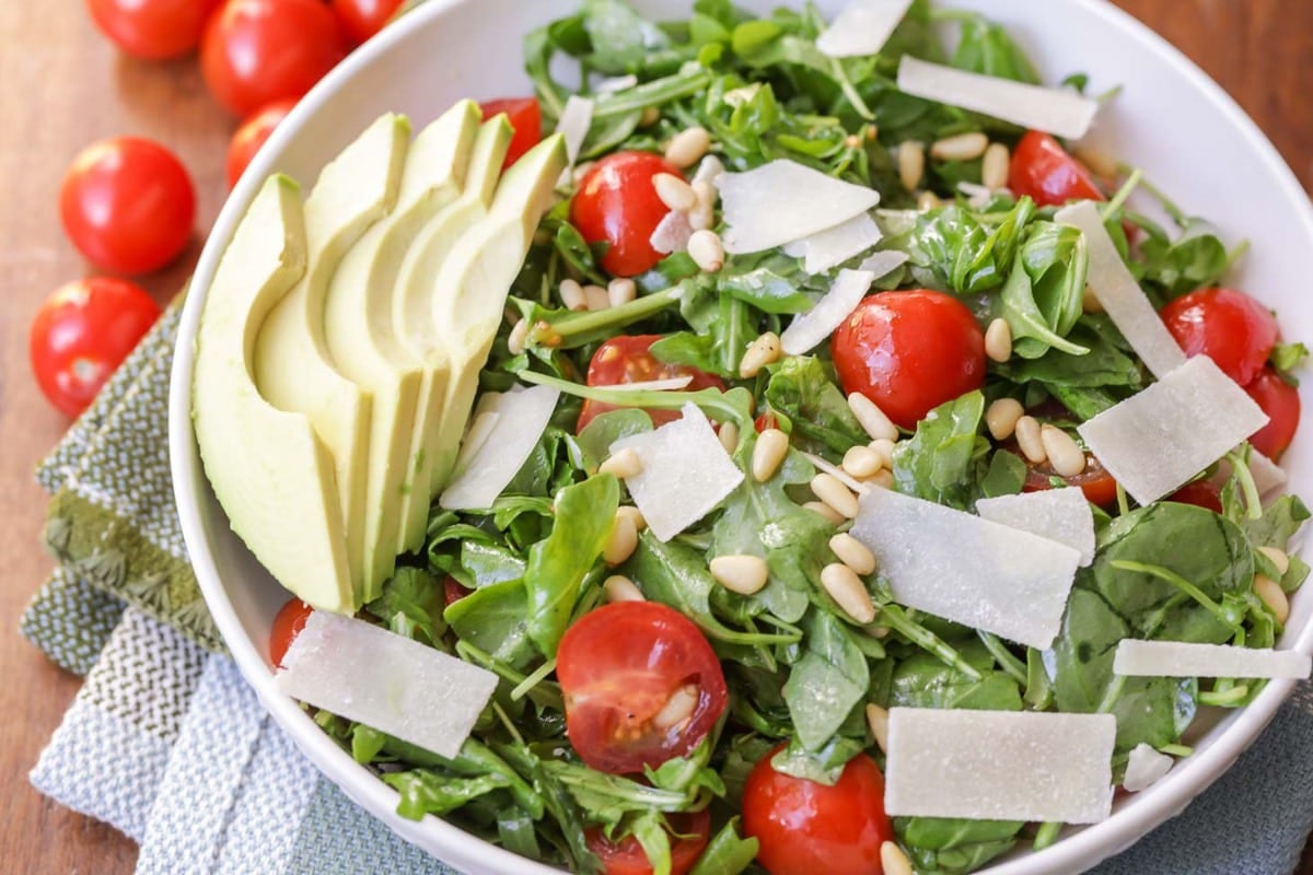 Healthy Dinner Ideas - Arugula salad topped with pine nuts, tomatoes, parmesan shavings and sliced avocados in a white bowl.