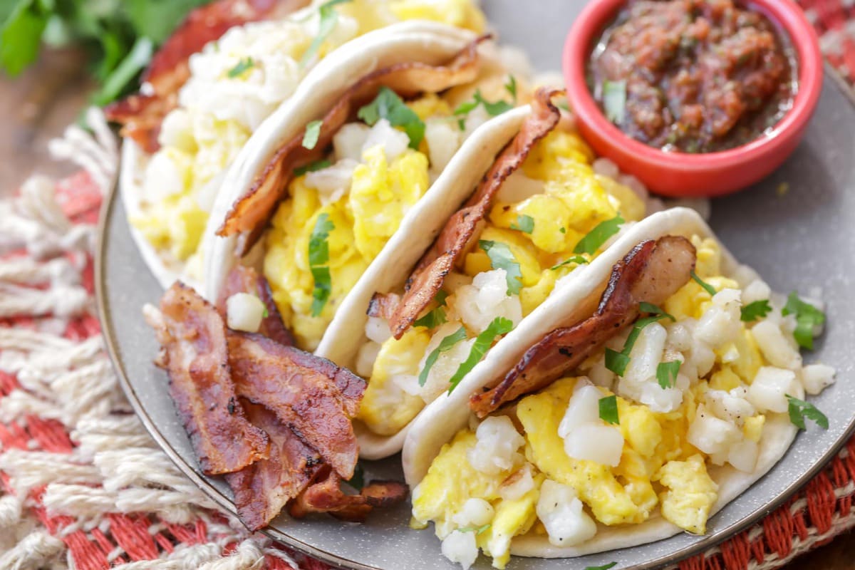 Easy Breakfast Ideas - Breakfast tacos garnished with cilantro with a side of bacon and a small bowl of red salsa on a white plate. 