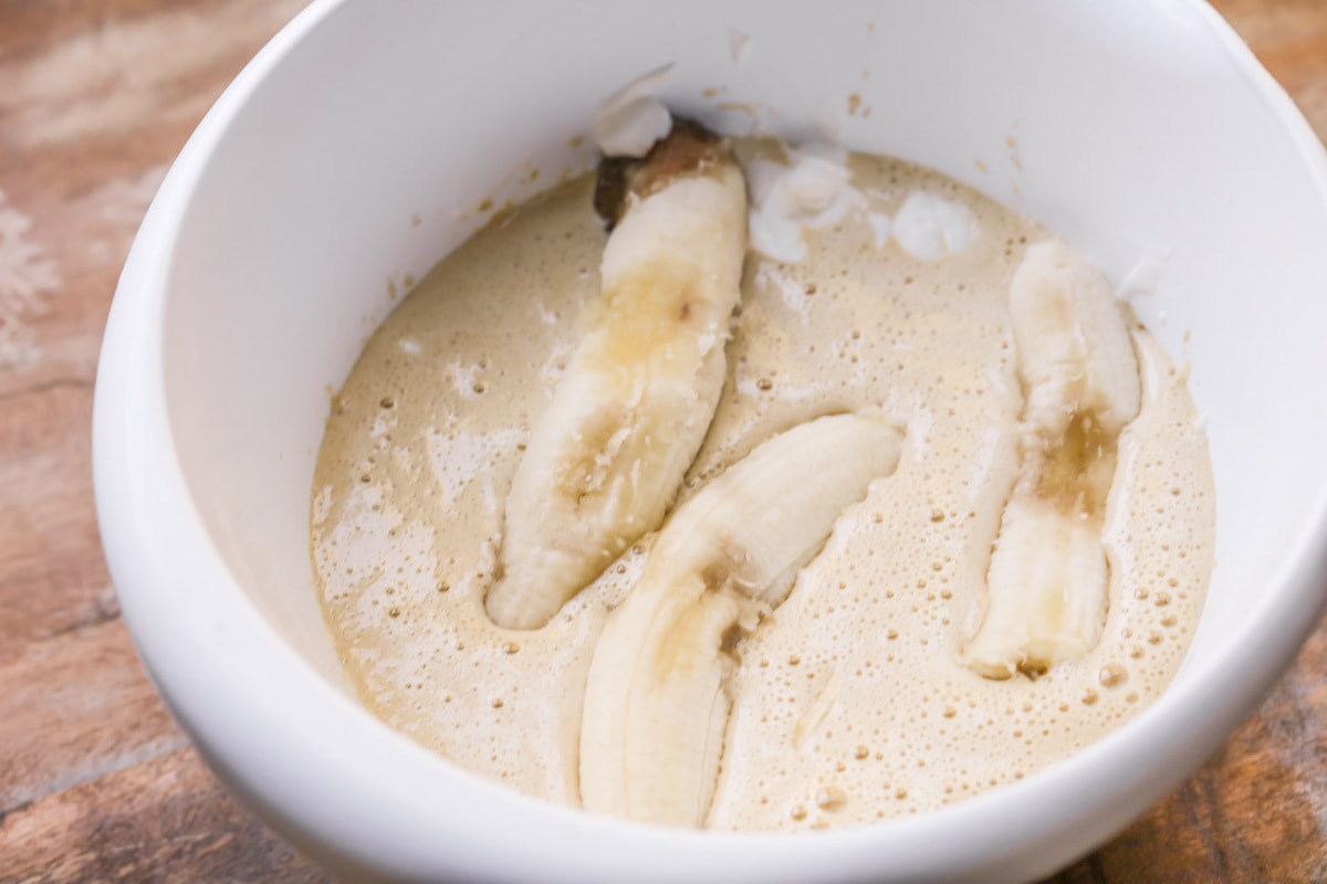 Adding ripe bananas to a batter in a white bowl.