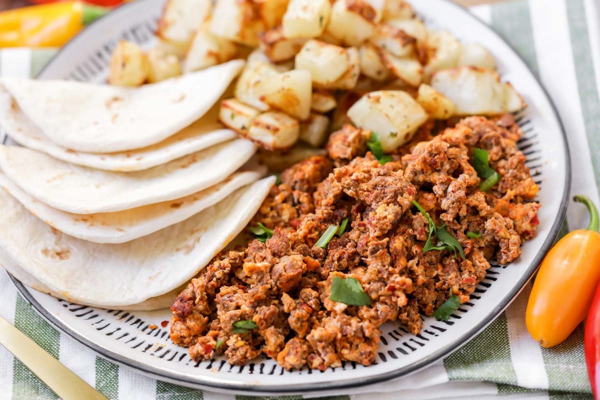 Easy Breakfast Ideas - chorizo and eggs with sides of folded flour tortillas and cubed potatoes on a blue and white plate. 
