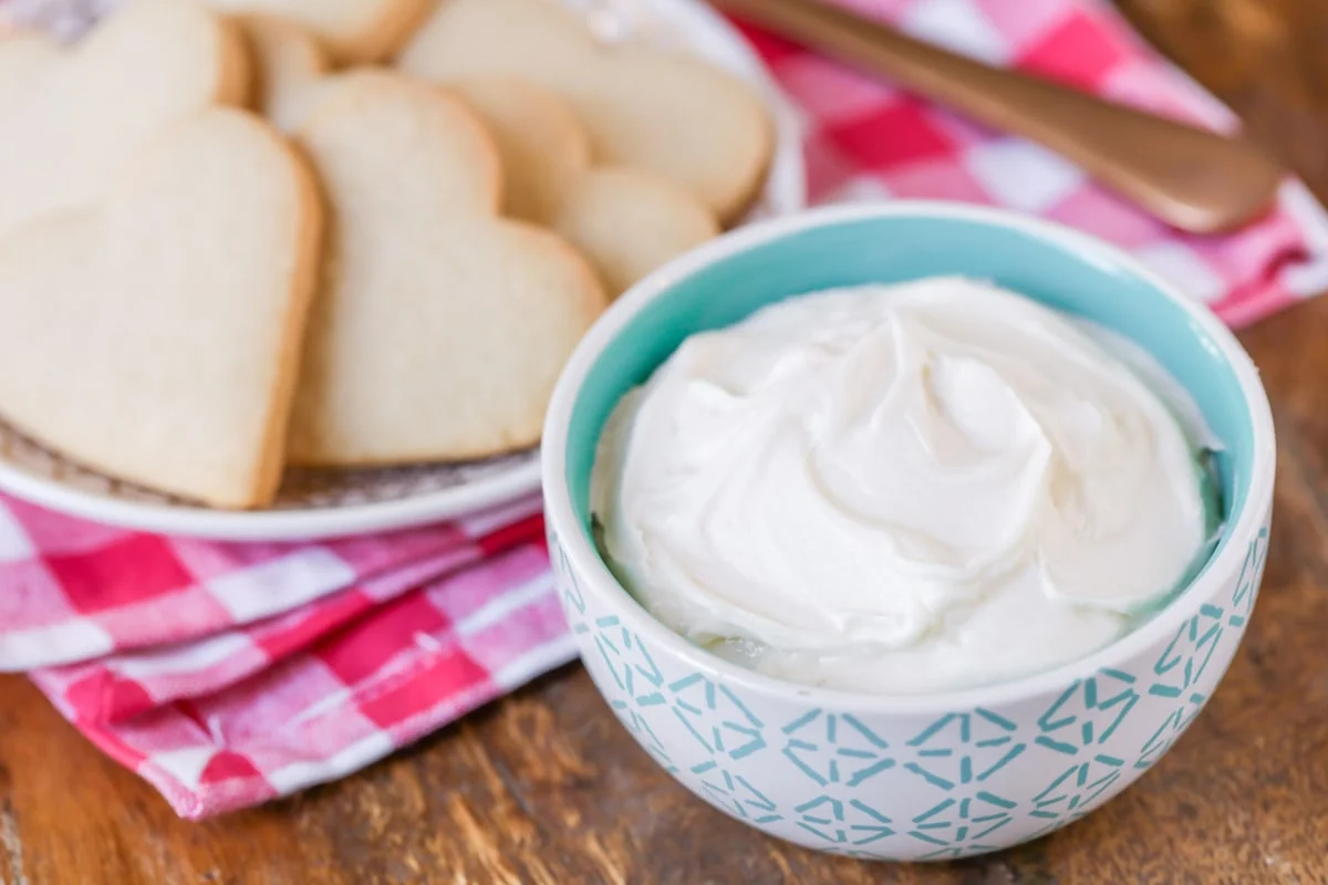 A bowl of sugar cookie frosting, with a plant of un-frosted, heart-shaped cookies in the background.