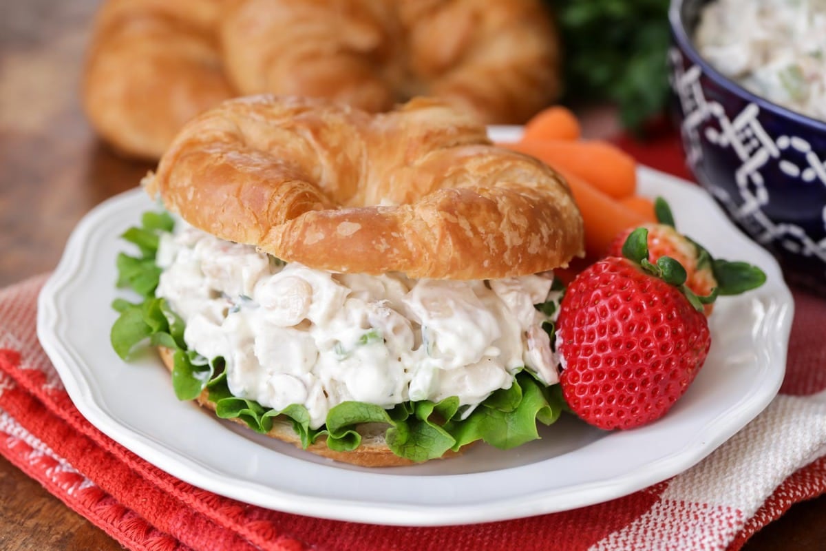 Chicken salad with lettuce on a croissant with a side of baby carrots and whole strawberries on a white plate. 