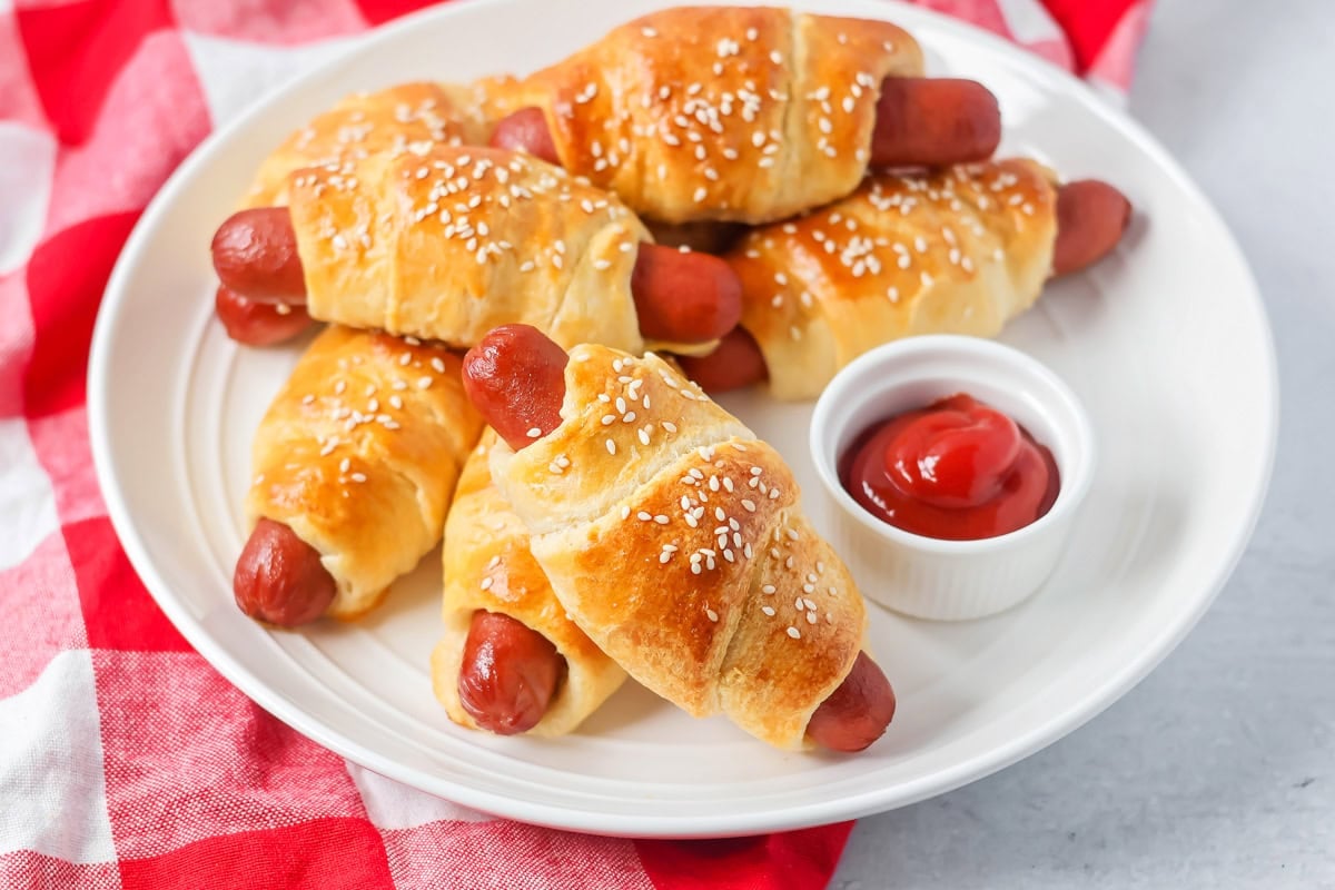 Pigs in a blanket stacked on each other on white plate served with ketchup.