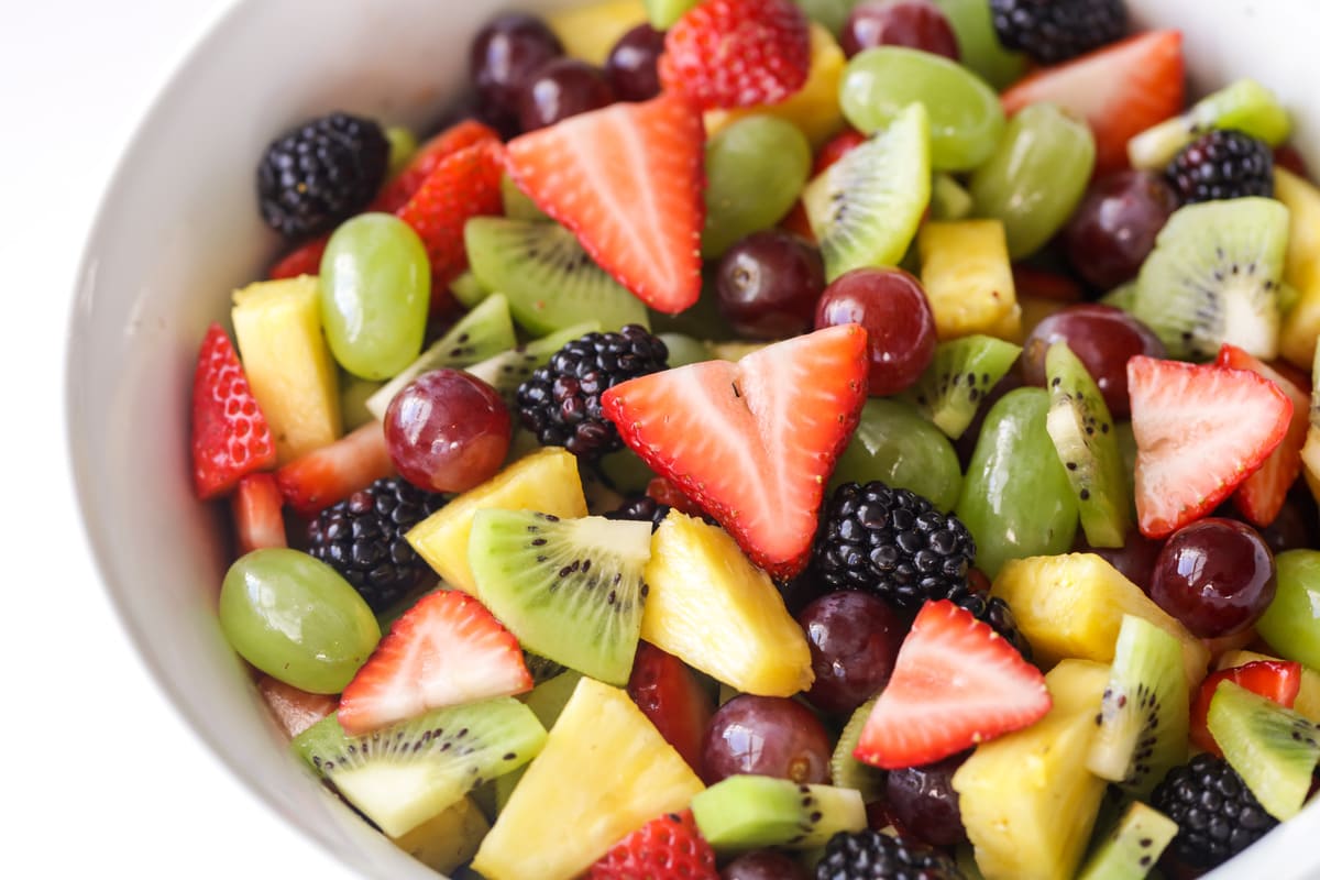 Fruit salad consisting of green grapes, red grapes, pineapple, kiwi, strawberries and blackberries in a white bowl. 