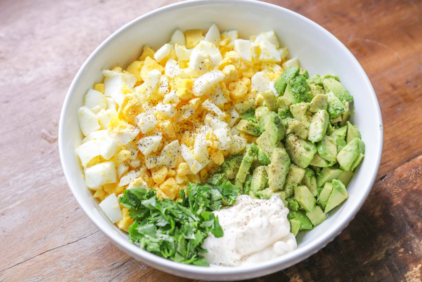 All ingredients for avocado egg salad in a white bowl on a wooden table. 