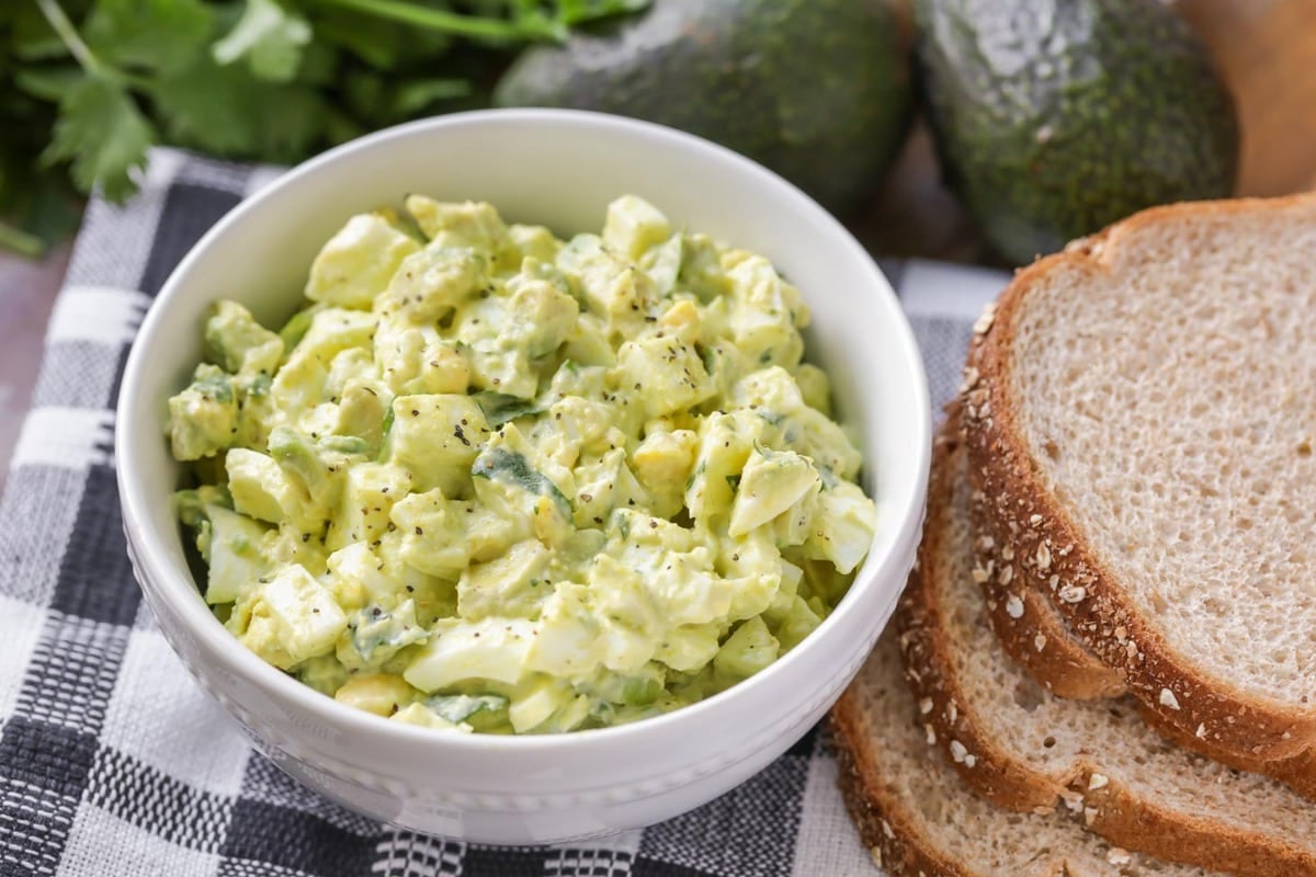 Avocado egg salad in a white bowl next to bread slices. 